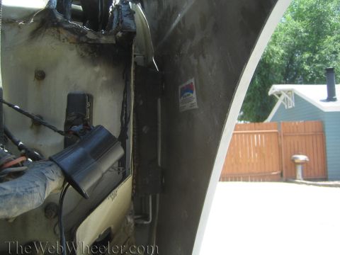 1990 Ford Bronco - Close-up inside a vehicle's fender well showing bare metal, wiring, and a backyard wood fence beyond.