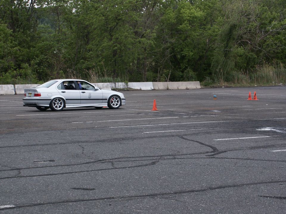 Silver BMW E36 sedan with aftermarket wheels driving on a paved track with orange cones