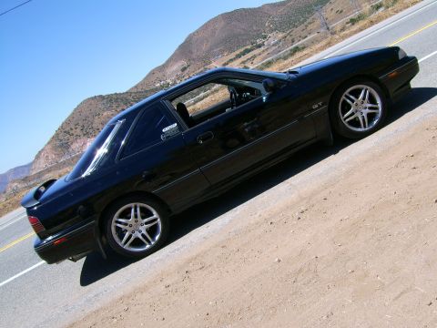 1989 Mazda MX-6 - A black 1989 Mazda MX-6 GT coupe is parked on the side of a road with a mountainous landscape in the background.