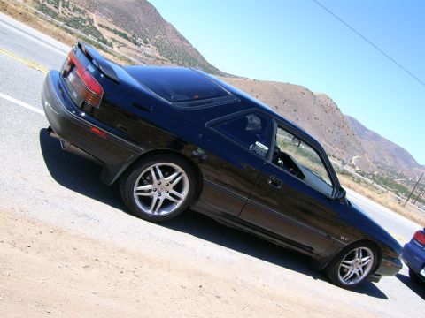 1989 Mazda MX-6 - A black 1989 Mazda MX-6 coupe is parked on the side of a road with mountains in the background.