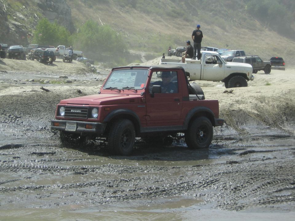 Red 1987 Suzuki Samurai driving through a muddy off-road course with other vehicles in the background