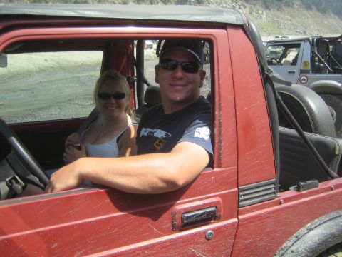 1987 Suzuki Samurai - A couple smiles from inside a red off-road vehicle, with another vehicle visible in the background.