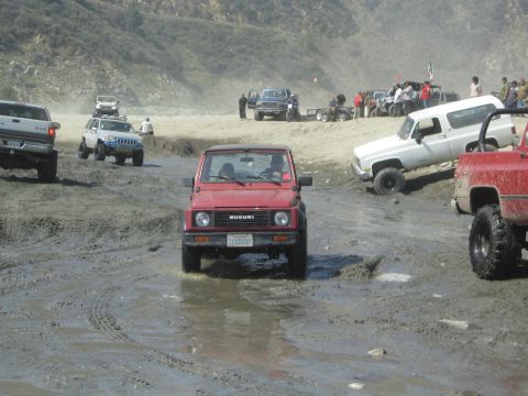 1987 Suzuki Samurai - Multiple off-road vehicles navigate a muddy terrain under a bright sky.