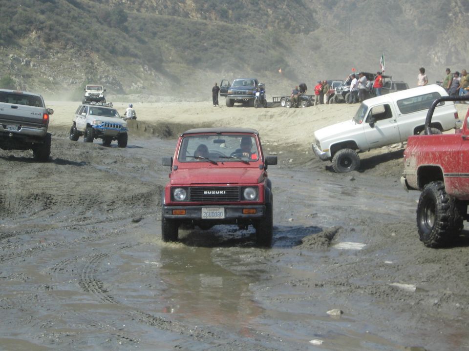 Red 1987 Suzuki Samurai driving through mud, with other off-road vehicles in the background