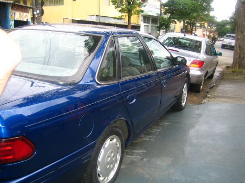 1995 Ford Taurus - A blue sedan is parked on a street next to another car. The vehicle is wet, possibly from rain.