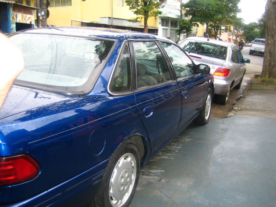 Blue sedan parked on street, another car behind it. Not a 1995 Ford Taurus