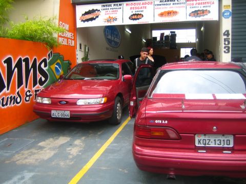 1995 Ford Taurus - Two red Ford sedans are parked side-by-side in front of a shop with services advertised on the wall.
