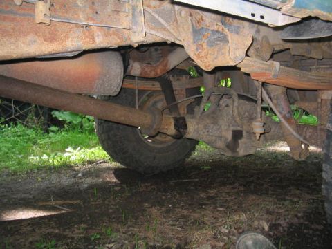 1979 Ford Bronco - Underside view of a vehicle's rear suspension showing a leaf spring, differential housing, driveshaft, and rusty frame components over dirt…