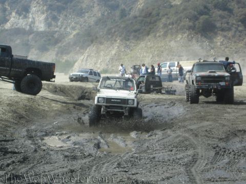 1987 Suzuki Samurai - Several off-road vehicles navigate a muddy terrain with spectators watching from the side.