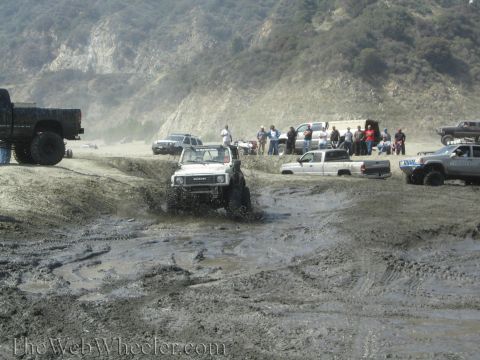 1987 Suzuki Samurai - Several off-road vehicles navigate a muddy terrain with spectators watching from the side.