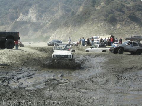 1987 Suzuki Samurai - A white 1987 Suzuki Samurai navigates a muddy obstacle course, with several other vehicles and onlookers present.