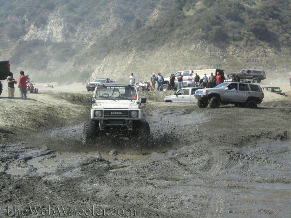 White 1987 Suzuki Samurai driving through mud, splashing water. Other vehicles and people in background
