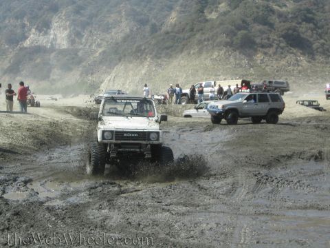 1987 Suzuki Samurai - A white 1987 Suzuki Samurai navigates a muddy off-road course with spectators in the background.