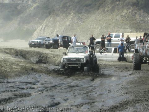 1987 Suzuki Samurai - A white 1987 Suzuki Samurai navigates a muddy terrain, surrounded by other off-road vehicles and onlookers enjoying the scene.