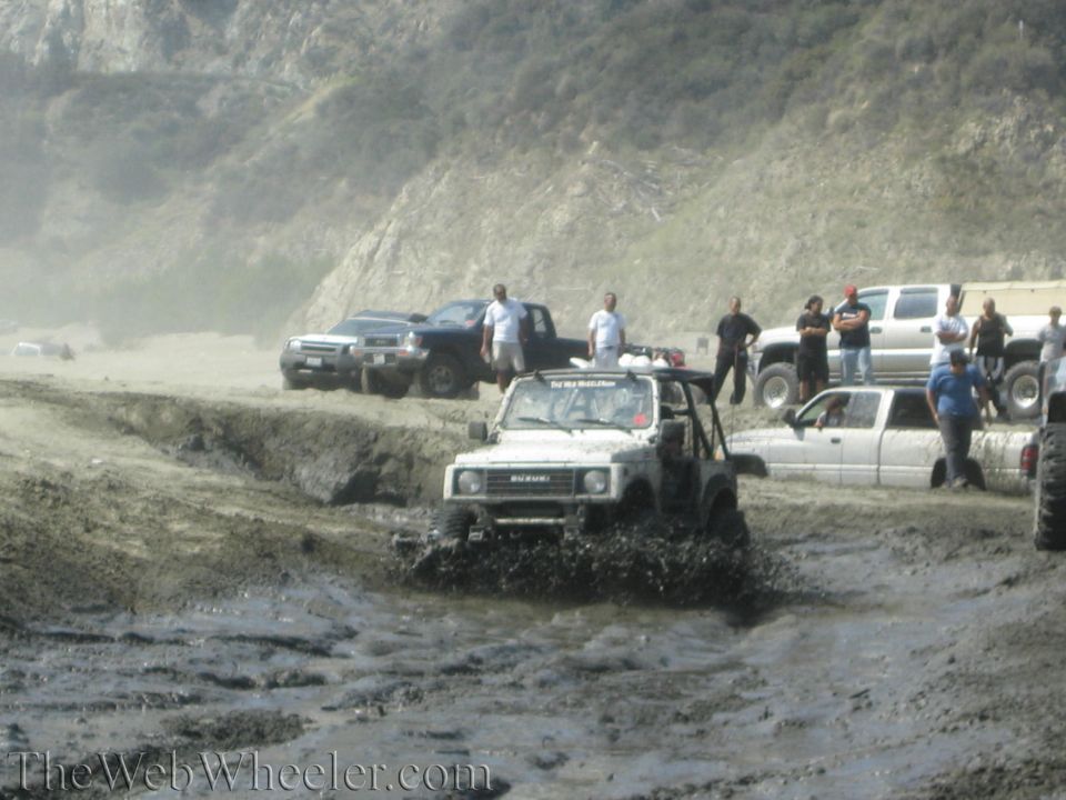 1987 Suzuki Samurai splashing through mud with other vehicles and people in the background on a beach