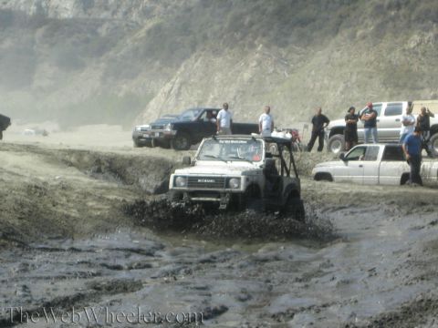 1987 Suzuki Samurai - Several off-road vehicles navigate a muddy terrain, with one Samurai prominently splashing through the mire.