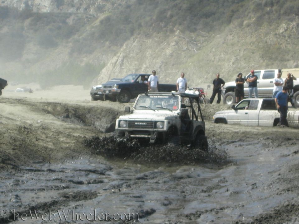White 1987 Suzuki Samurai driving through mud with other vehicles nearby