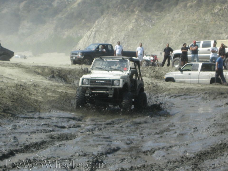White Suzuki Samurai with lifted suspension driving through mud, front view. "The Web Wheeler.com" watermark visible
