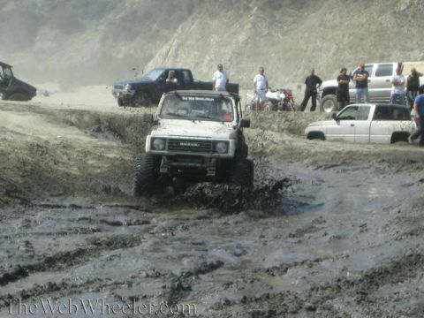 1987 Suzuki Samurai - A white 1987 Suzuki Samurai navigates a muddy off-road course with other vehicles and spectators present.