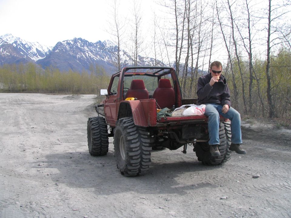 Guy sitting on the back of a red off-road vehicle with huge tires, mountains in background