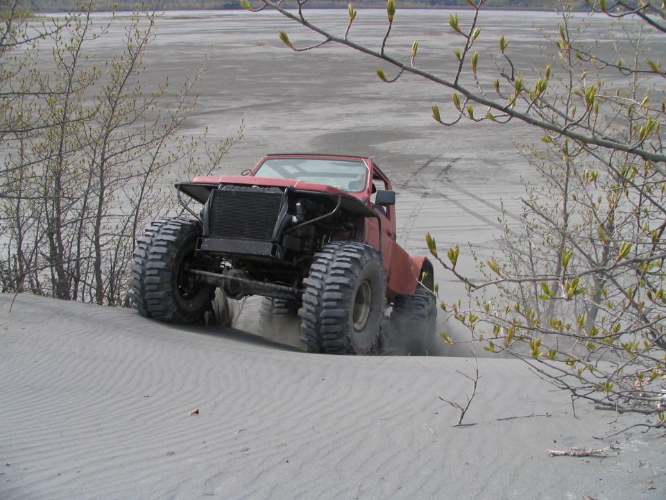 Red off-road vehicle with large tires driving on sand, front grille removed