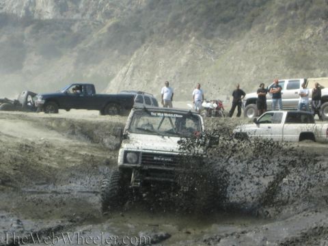 1987 Suzuki Samurai - A white 1987 Suzuki Samurai navigates a muddy obstacle, sending up a spray of water and dirt.