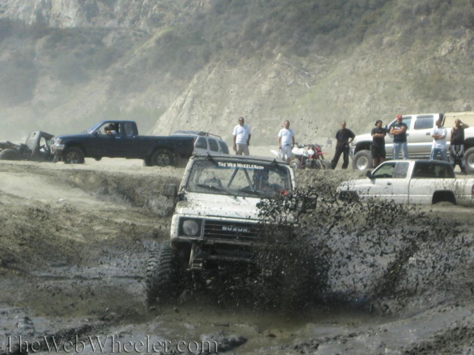 White 1987 Suzuki Samurai splashing mud, front view, with other vehicles and people in the background