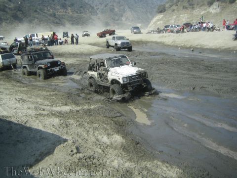 1987 Suzuki Samurai - Several off-road vehicles are seen driving through a muddy terrain, with hills and spectators in the background.
