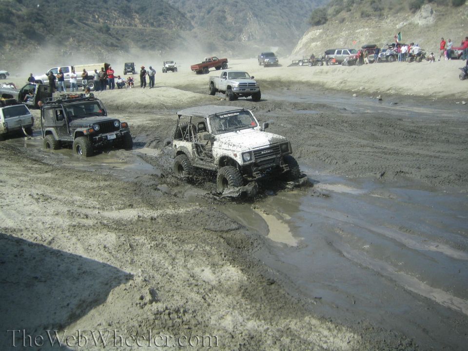 White Suzuki Samurai navigating a muddy track, with other off-road vehicles and spectators in the background