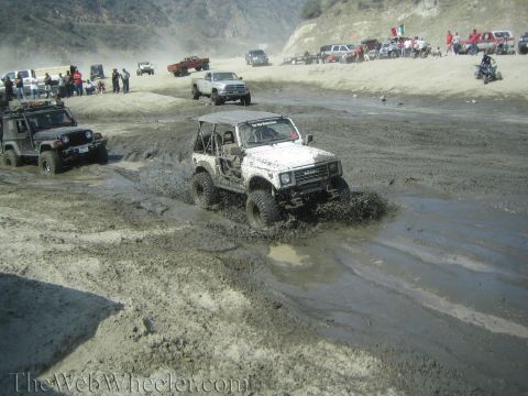 1987 Suzuki Samurai - A white 1987 Suzuki Samurai navigates a muddy off-road course, kicking up water and dirt.