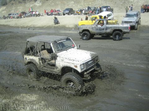 1987 Suzuki Samurai - A 1987 Suzuki Samurai navigates a muddy terrain, with several other vehicles and spectators visible on a nearby embankment.