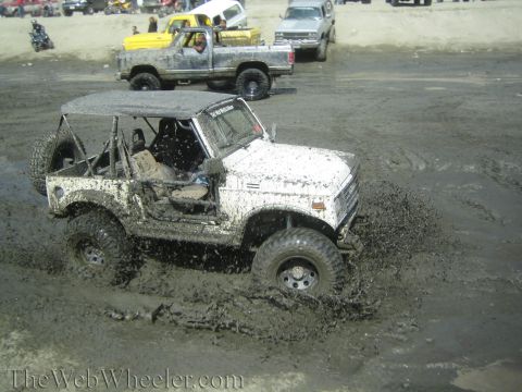 1987 Suzuki Samurai - A white 1987 Suzuki Samurai navigates a muddy terrain, kicking up spray as it goes.