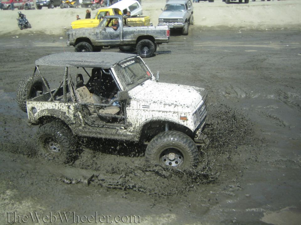 White 1987 Suzuki Samurai with roll cage driving through mud, tires deep in muck