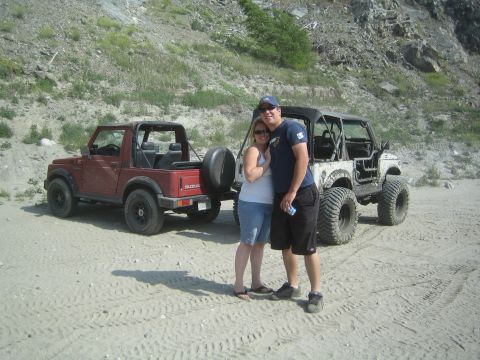 1987 Suzuki Samurai - A couple poses in front of a red 1987 Suzuki Samurai and a heavily modified white Jeep on a sandy, hilly terrain.
