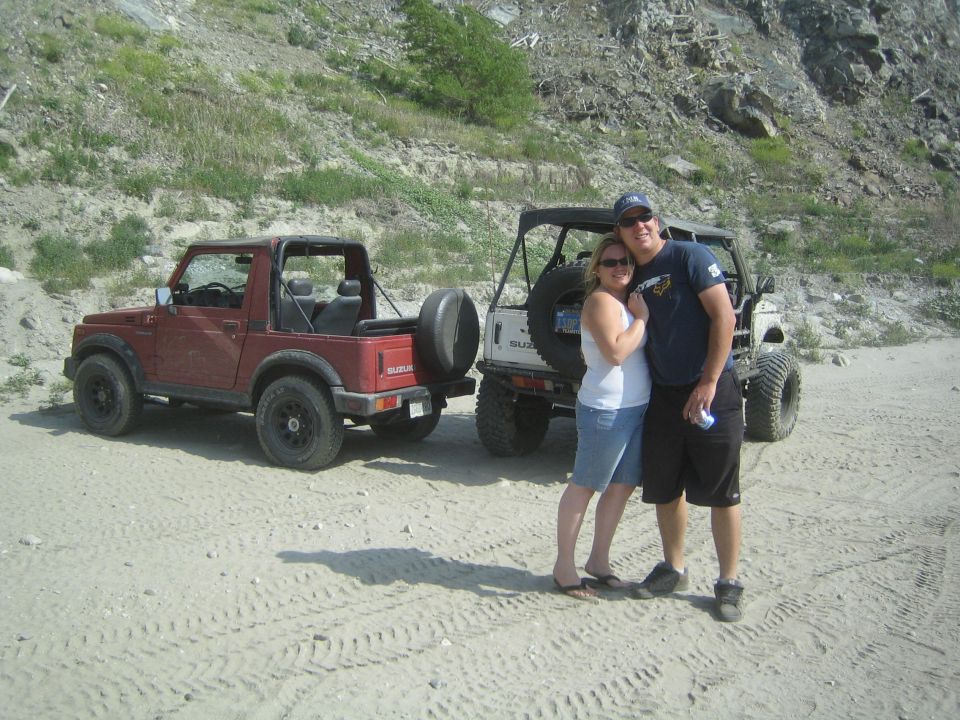 Couple posing in front of two red and white Suzuki Samurai vehicles on a sandy, rocky terrain