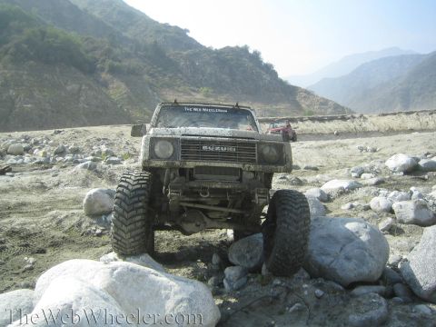 1987 Suzuki Samurai - Mud-covered 1987 Suzuki Samurai navigating a rocky terrain with a second vehicle visible in the distance.