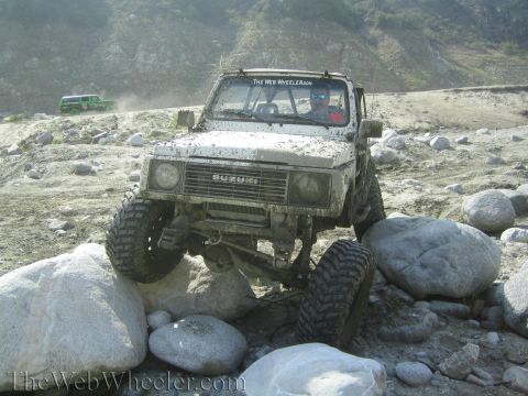 1987 Suzuki Samurai - A white 1987 Suzuki Samurai is shown off-roading over large rocks, with another green vehicle in the background.