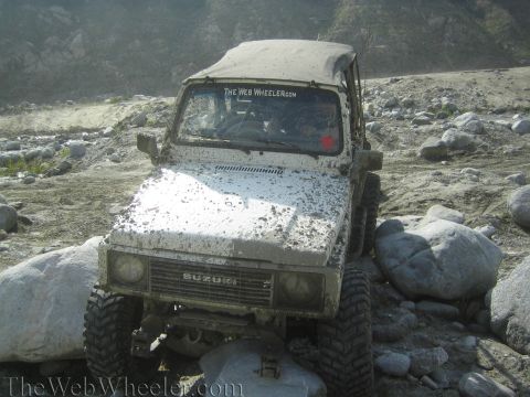 1987 Suzuki Samurai - This Suzuki Samurai is shown off-roading through a rocky, dry riverbed, its white body splattered with mud.