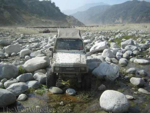 1987 Suzuki Samurai - A white 1987 Suzuki Samurai navigates a rocky riverbed with mountains in the background.