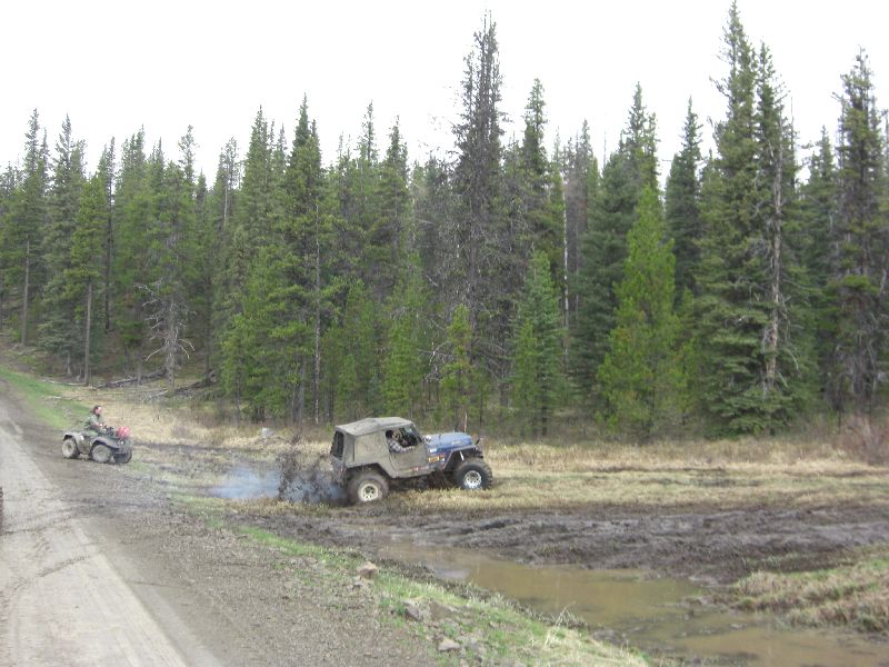 Jeep is stuck in mud, an ATV is on a dirt road nearby, surrounded by trees