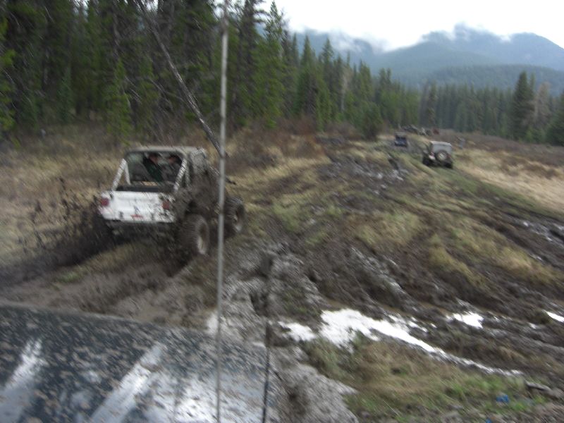 Muddy off-road vehicles navigating a steep, wet, and uneven hill with trees and mountains in the background