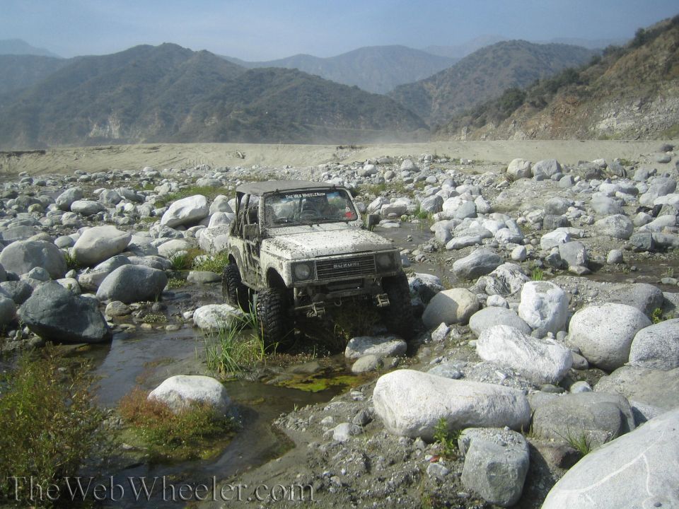 1987 Suzuki Samurai navigating a rocky riverbed with mountains in the background