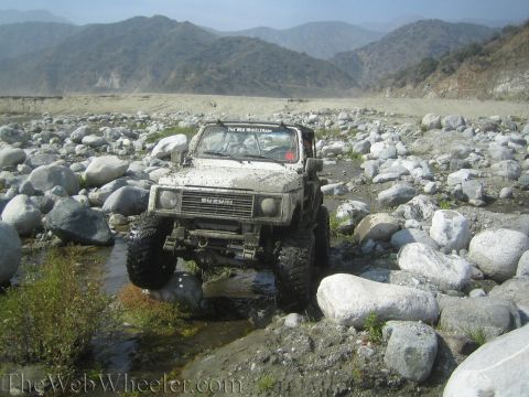 1987 Suzuki Samurai - A Suzuki Samurai navigates a challenging, rocky terrain in a dry riverbed, with mountains visible in the distance.