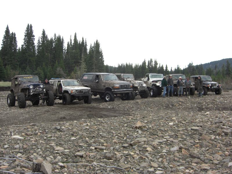 Several modified off-road vehicles parked on a rocky surface with trees in the background. People stand near some of the vehicles