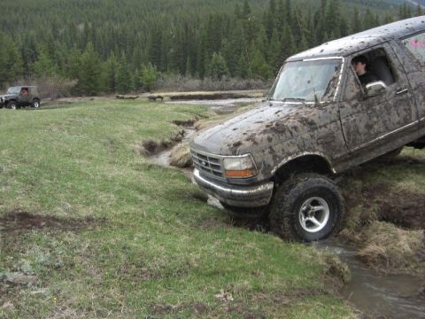 1995 Ford Bronco - A muddy 1995 Ford Bronco navigates a stream during an off-road excursion, with another SUV visible in the distance.