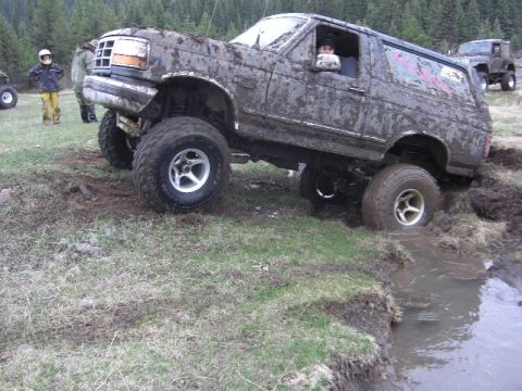 1995 Ford Bronco - A heavily muddied SUV is shown off-roading, with one wheel deep in a water-filled rut.