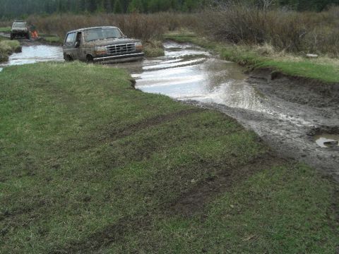 1995 Ford Bronco - Two SUVs navigate a muddy, water-filled trail through brush and grass.
