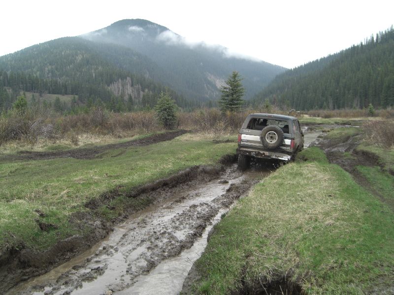 1995 Ford Bronco driving through mud on a dirt road with mountains in the background