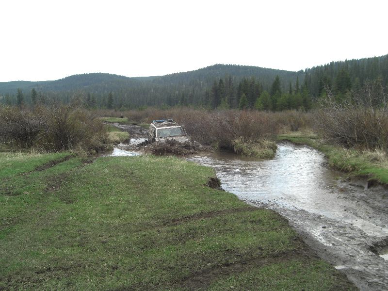 off-road vehicle stuck in mud and water with trees and hills in background