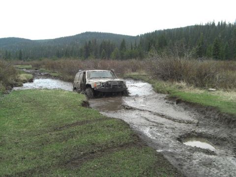1995 Ford Bronco - An off-road vehicle navigates a challenging muddy terrain with water and dense foliage.